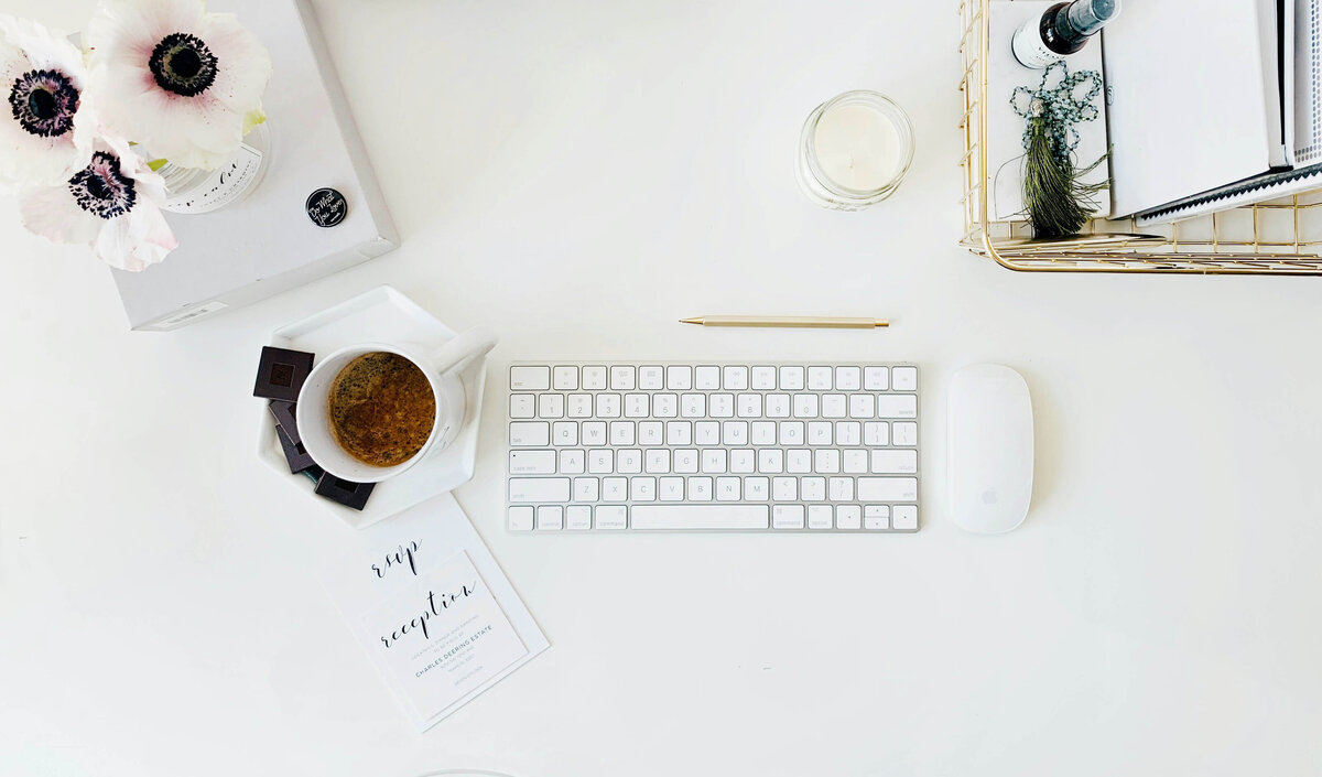 Flatlay of a wedding photographer’s desk with flowers, coffee, chocolate, a keyboard, and a notebook basket, symbolizing workflow organization and productivity.