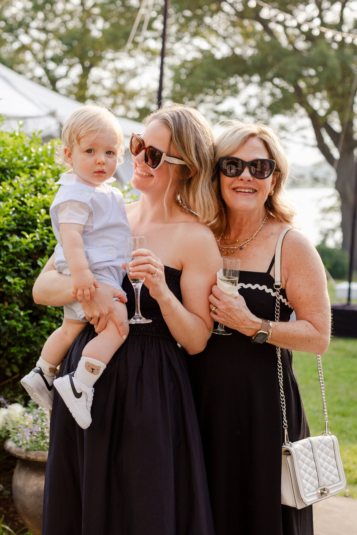 two women holding a baby with glass during cocktail hour of the outdoor rehearsal dinner at Wainwright House
