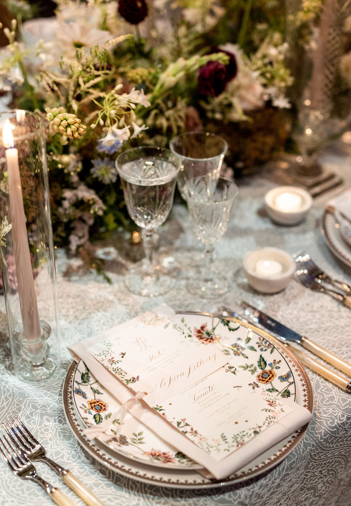 This is where the photo was taken at Washington D.C., Washington National Cathedral by photographer Twah Dougherty. The elegantly set dining table features intricate floral-patterned plates, crystal glasses, and menu cards. The table is decorated with a tall lit candle, small votive candles, and lush floral arrangements, creating a sophisticated and romantic ambiance. Current Films also captured this beautiful setting as videographers.