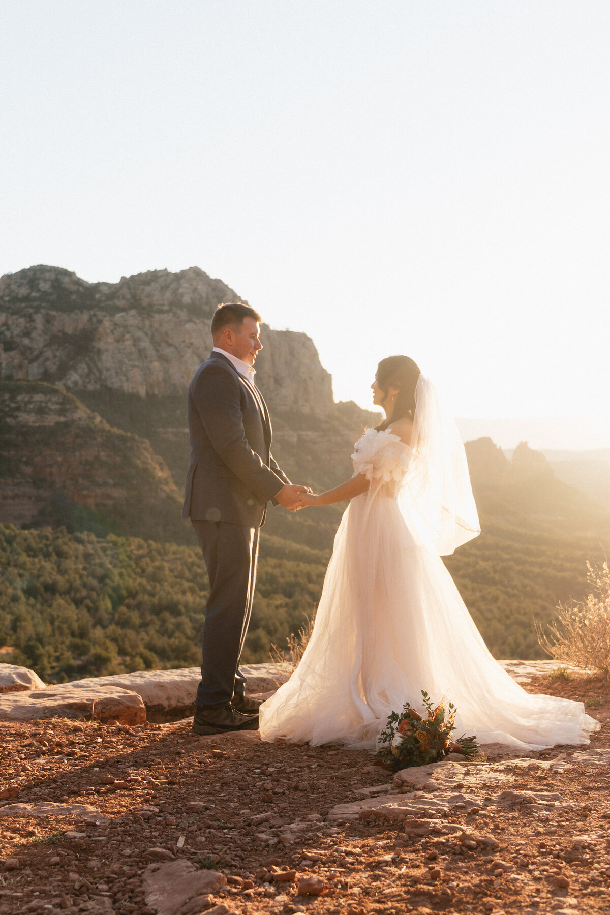 Bride and groom holding hands during their sunset elopement ceremony at Merry Go Round Rock in Sedona, Arizona, with dramatic cliffs and golden light in the background.