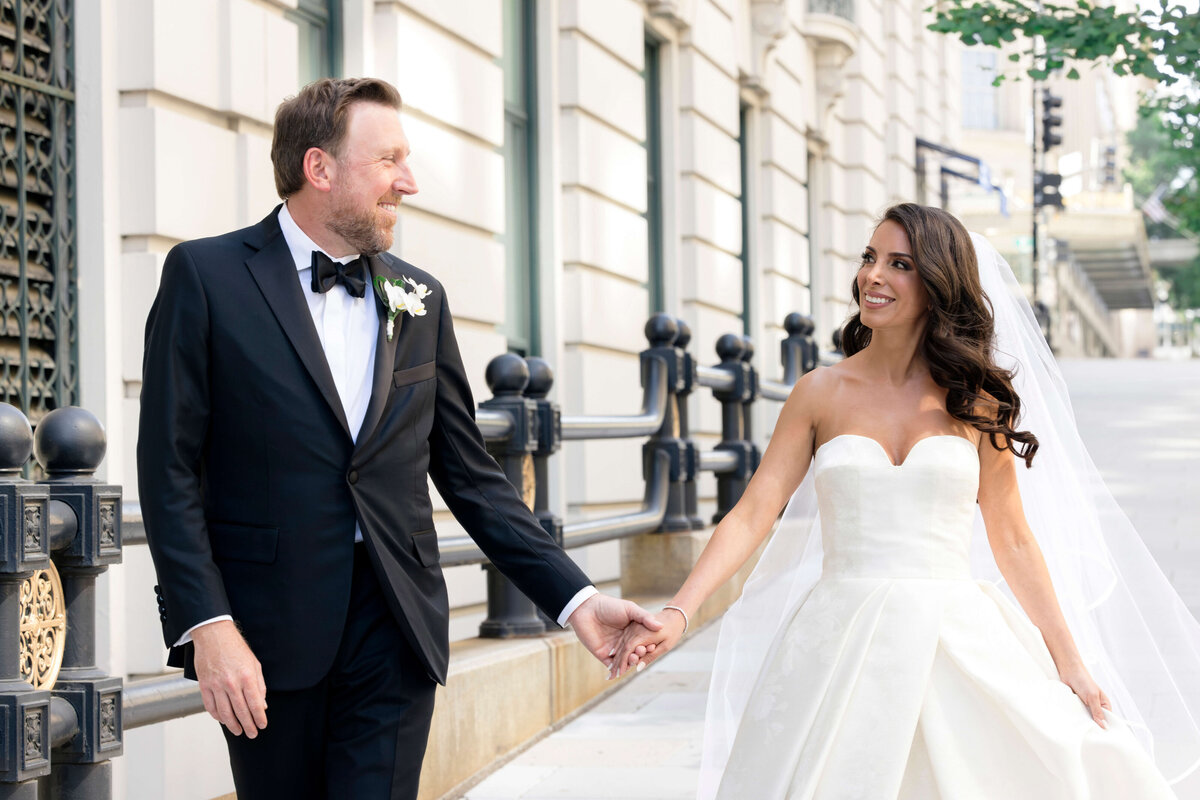 A bride in a strapless white gown and veil holds hands with a groom in a black tuxedo and bow tie. They walk together on a city sidewalk, smiling at each other, with a building and iron railing in the background.