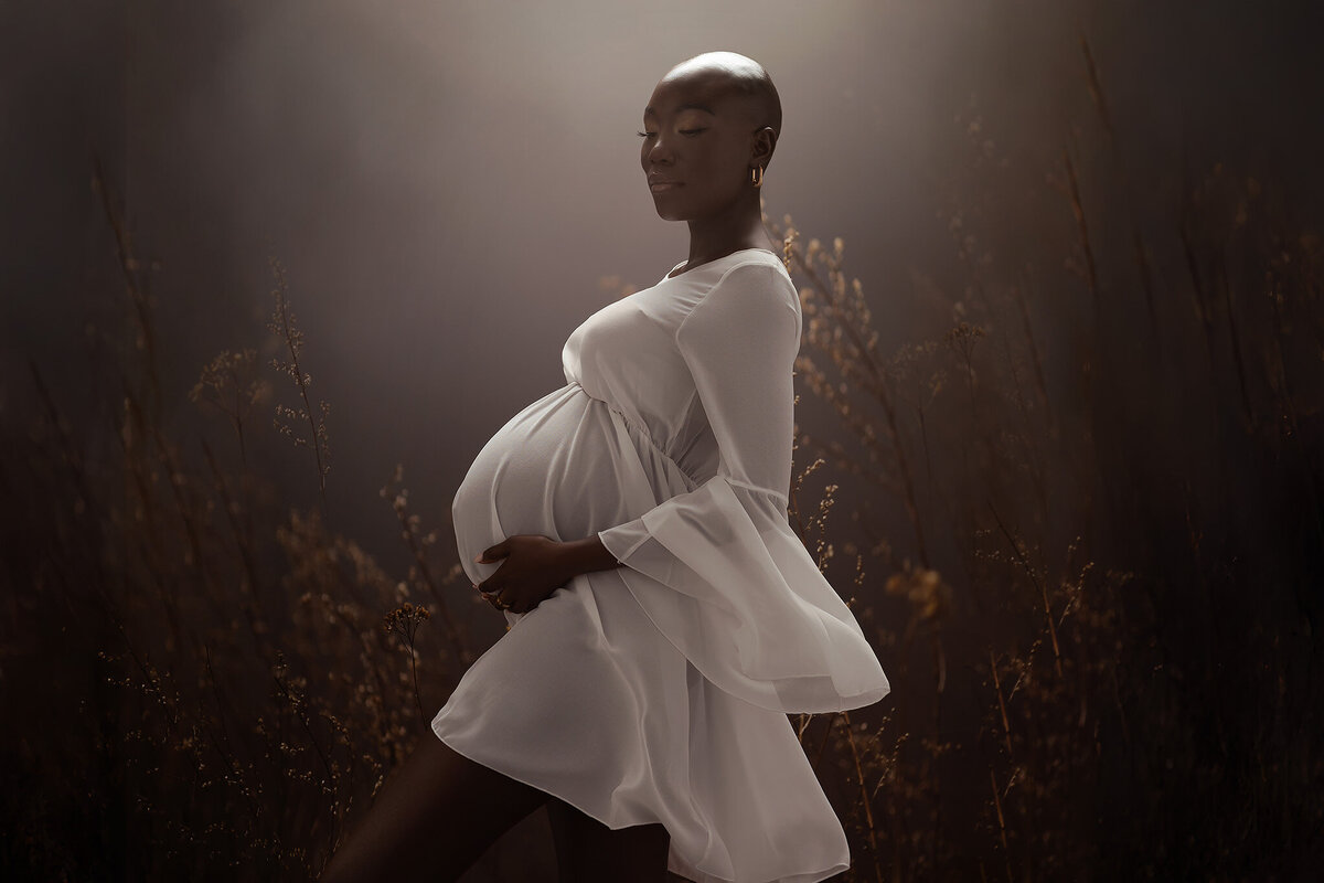 A serene woman stands gracefully in a flowing white dress, cradling her belly as golden light filters through tall, delicate grasses. The peaceful, dreamy atmosphere of this maternity photograph evokes the beauty of motherhood and nature.