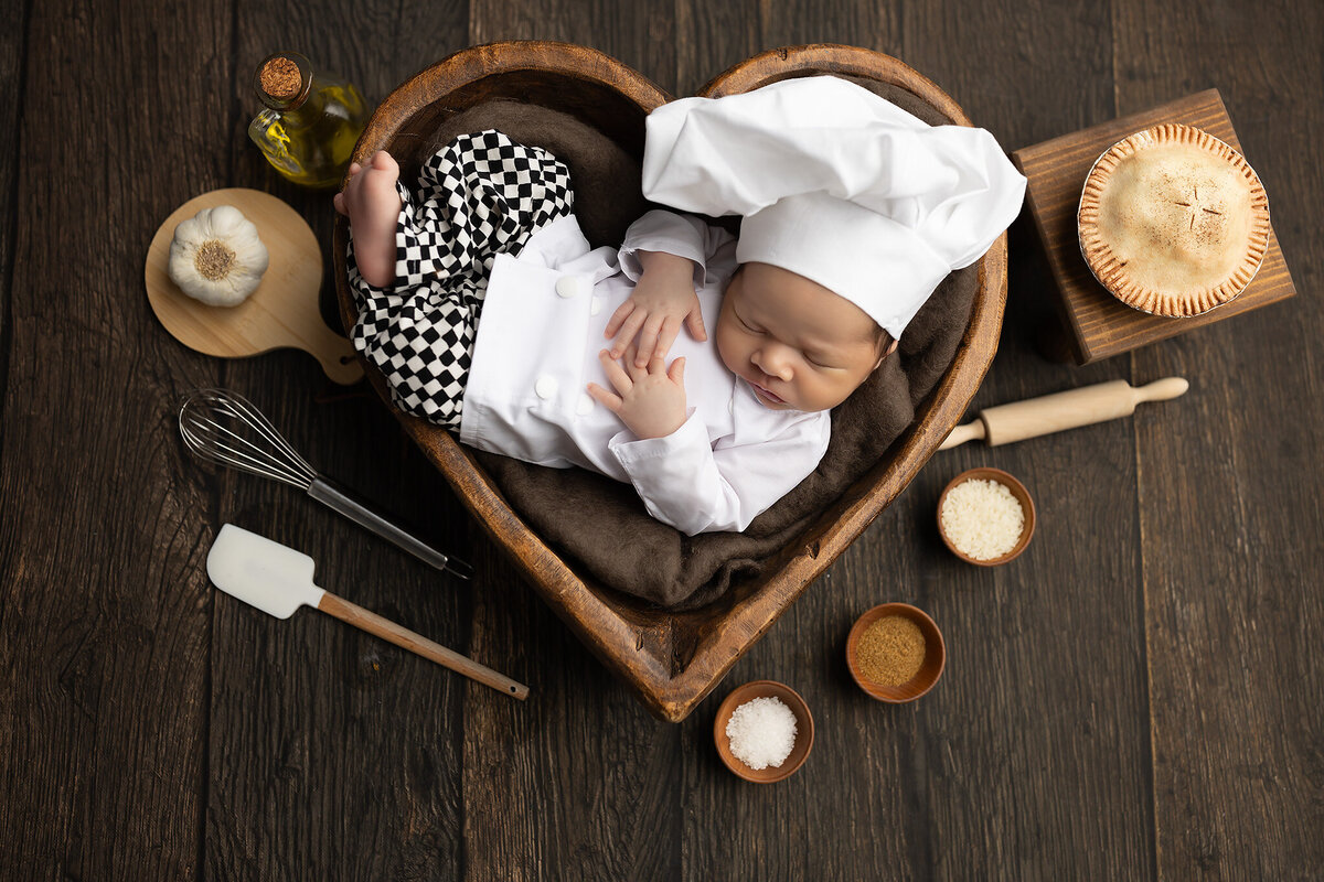 Baby in chef outfit lying in a heart-shaped bowl surrounded by kitchen tools, garlic, olive oil, and ingredients – Colleyville baby photography studio