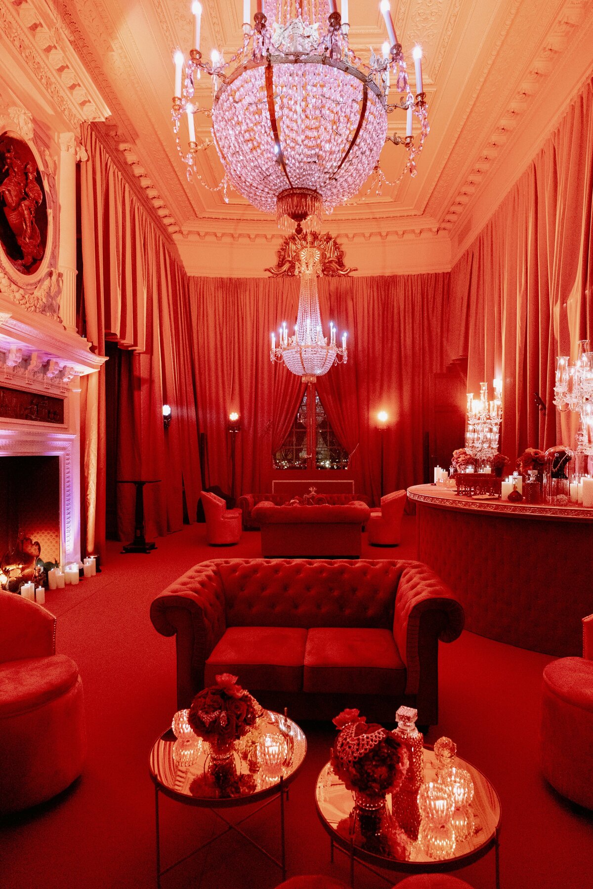 The photo was taken at the Shangri La Hotel & Ritz Paris Hotel in Paris, France, by the talented photographer Matteo Coltro, with Zen Film Works as Videographers. A lavish, dimly lit room decorated entirely in red is featured in the image. The room showcases plush velvet couches, elegant crystal chandeliers hanging from a high ornate ceiling, and draped curtains. The cozy ambiance is enhanced by small tables with floral arrangements and candlelit lamps, creating a truly luxurious setting.
