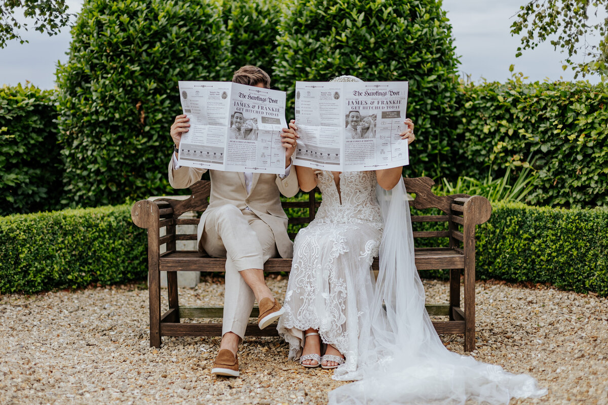Bride and groom sat on a bench in a garden holding newspapers infront of their faces
