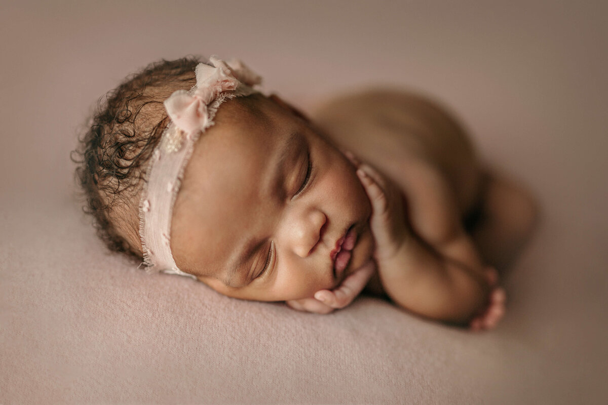newborn baby girl holding her cheeks wearing a pink headband with bows on a pink backdrop in a newborn session