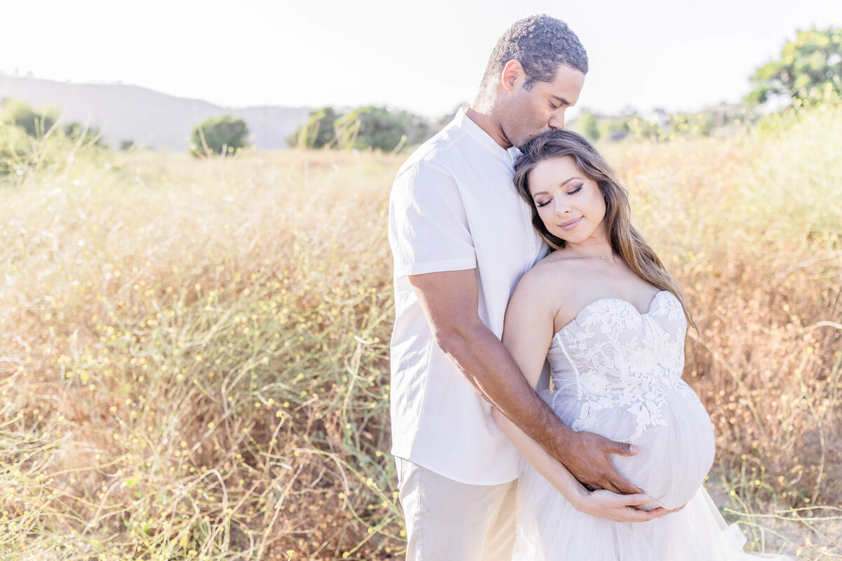 A couple stands in a sunlit field, embracing. The man gently kisses the womans forehead while she cradles her pregnant belly. They are surrounded by tall, golden grasses under a clear sky.