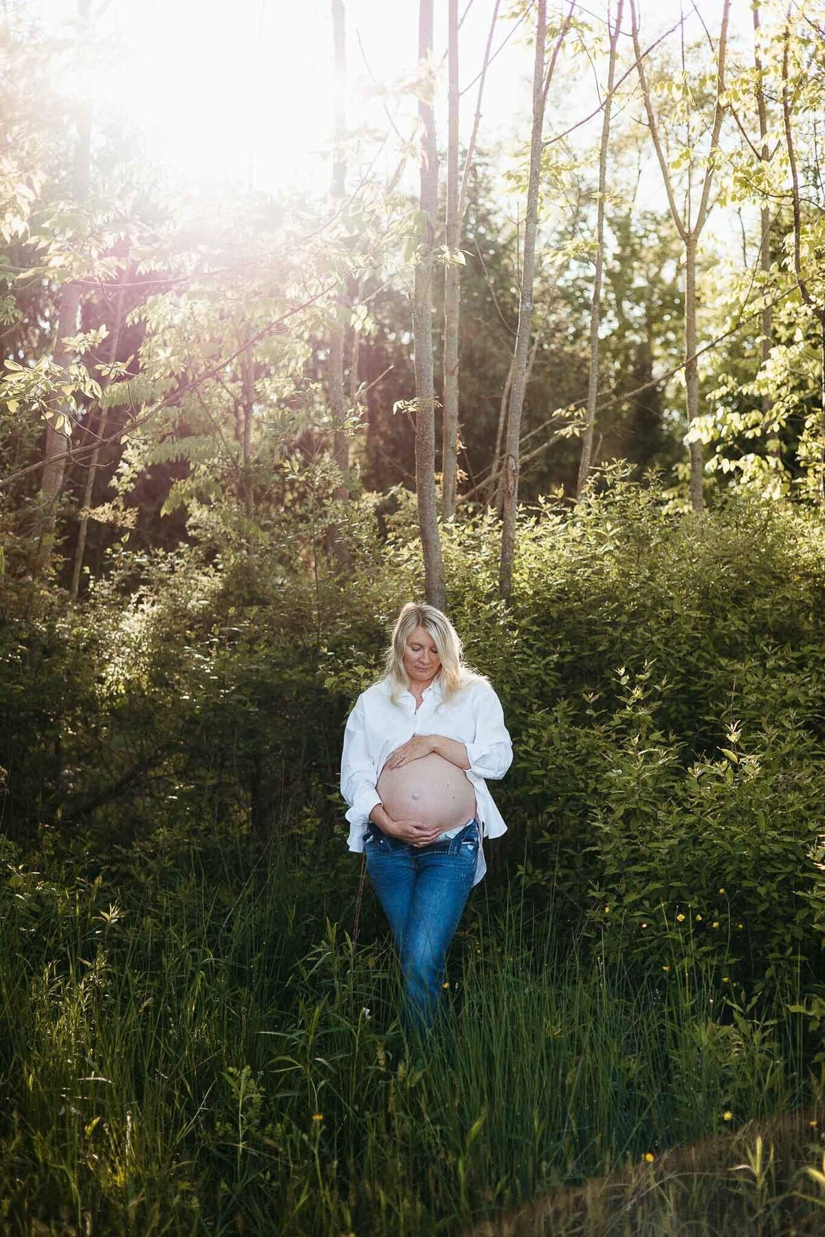 A natural maternity photo in a Cleveland woodland, featuring a pregnant mother in casual jeans and a white shirt cradling her belly.
