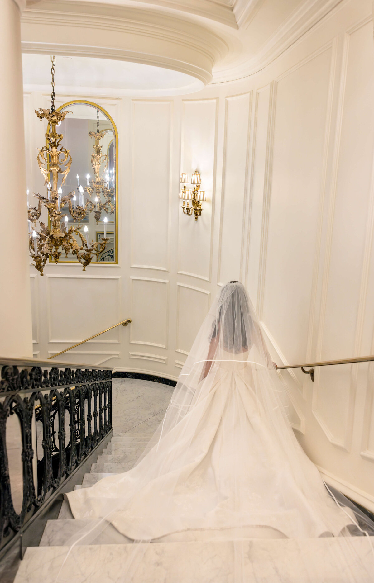 A bride in a long white gown and veil descends a grand staircase with ornate railings. A large, gold-framed mirror and elegant chandelier hang on the wall, enhancing the luxurious atmosphere.