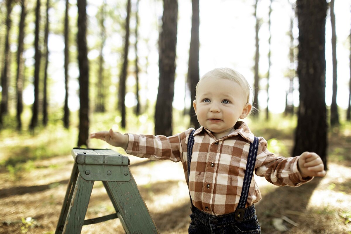 Birthday boy in pine trees, standing on a mini ladder, with a bright smile and his hands resting on the ladder