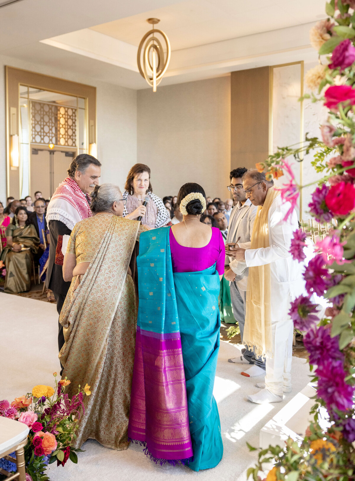 A group of people stands in a brightly decorated room during a ceremony. A woman in a blue and purple sari is prominent, accompanied by others in traditional attire, surrounded by colorful flowers and a seated audience in the background.