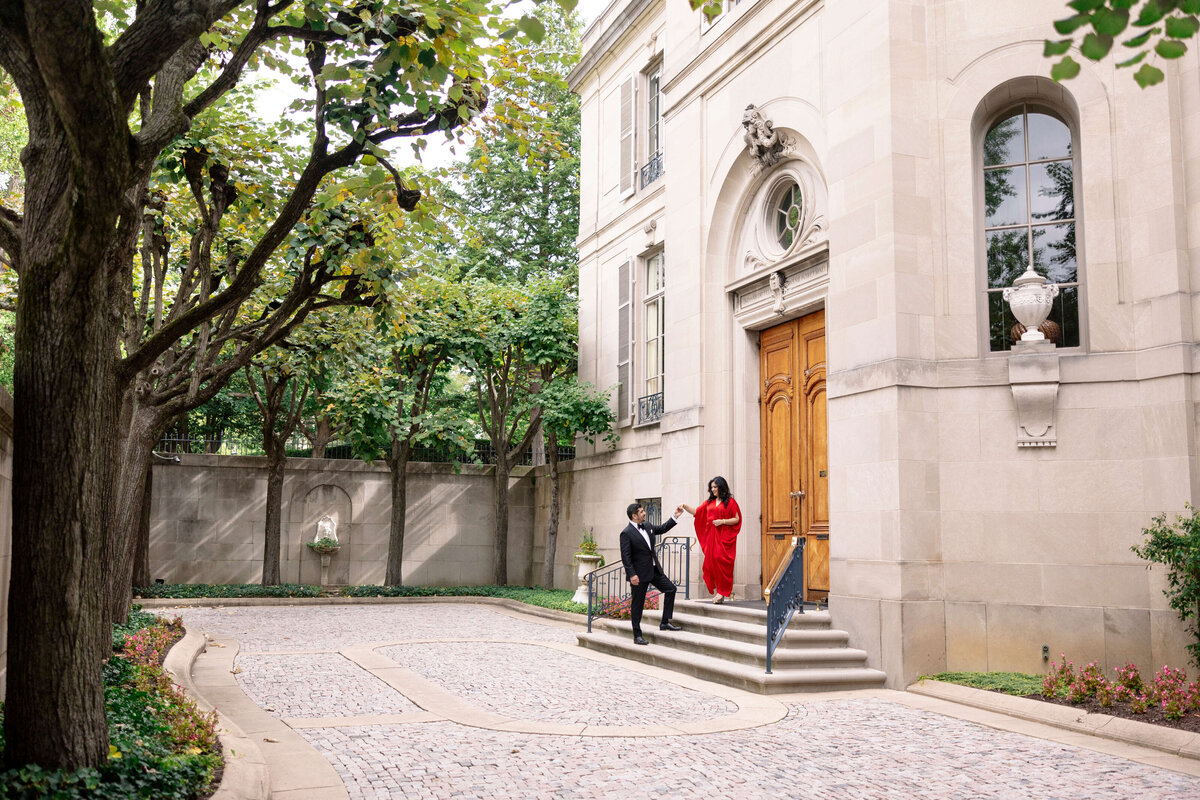 A couple stands on the steps of an elegant stone building with large wooden doors. The woman in a red dress and the man in a black suit are under a canopy of trees, with a cobblestone path leading up to the entrance.