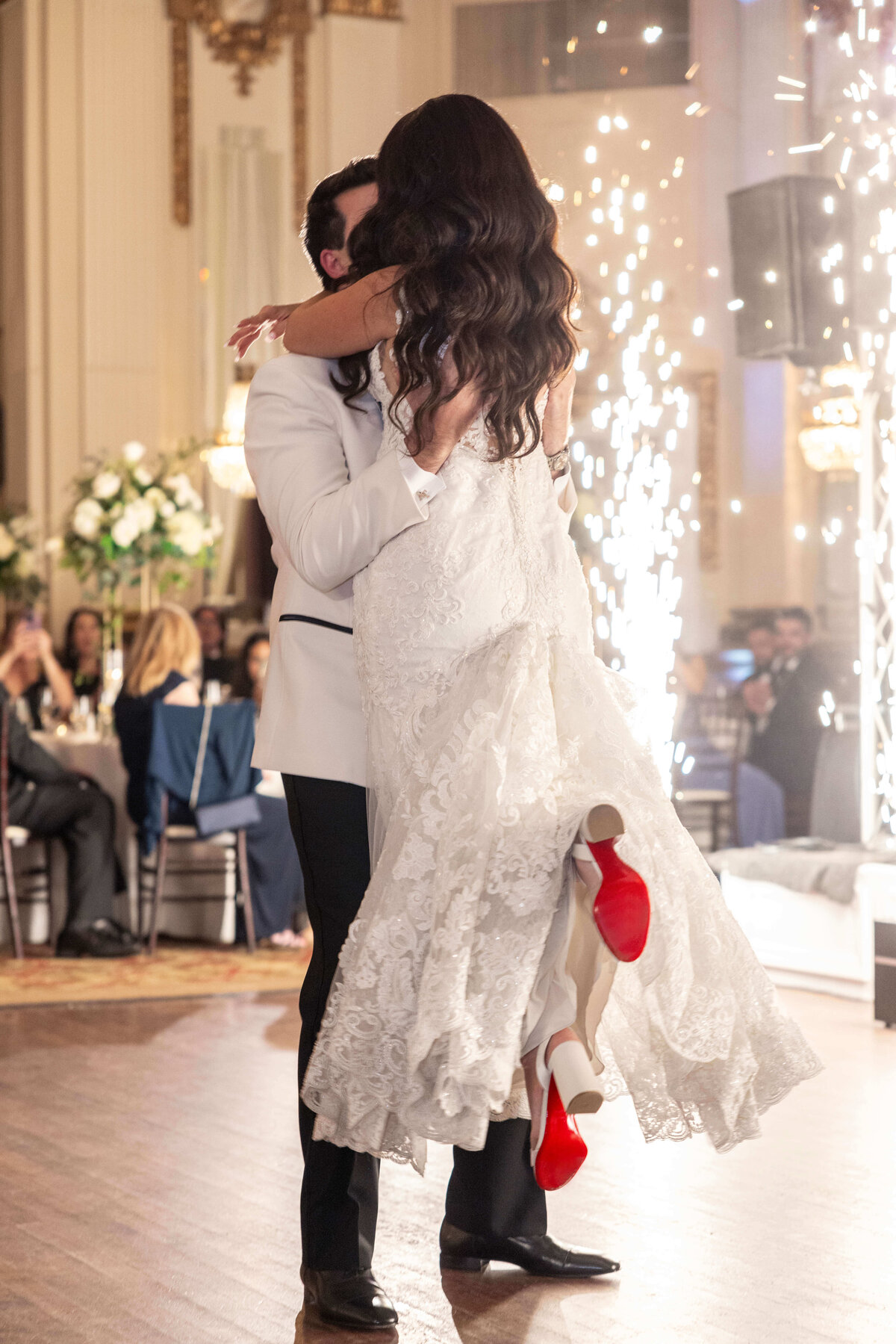 A groom in a white suit lifts the bride, who is wearing a white lace dress and red-soled shoes, as they share a joyful moment on the dance floor. Sparkling fireworks and elegantly decorated tables are in the background.