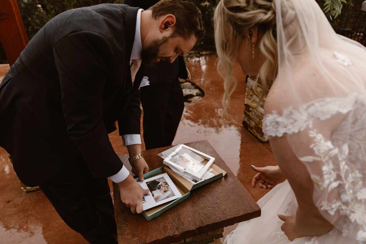 Bride and groom open a box of printed wedding photos on a rainy ceremony day, sharing a quiet and emotional moment over captured memories.