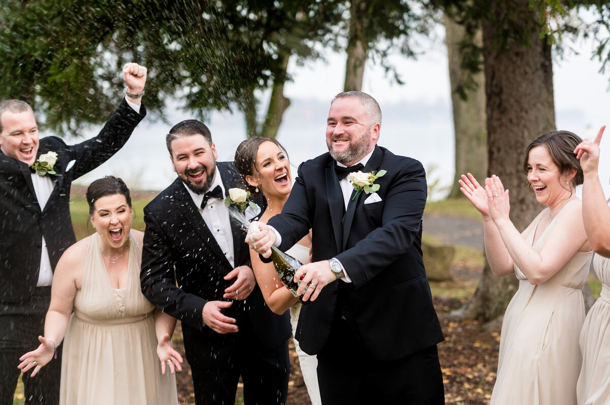 Groom wearing a tuxedo spraying a bottle of champagne as his wife and bridal party cheer him on