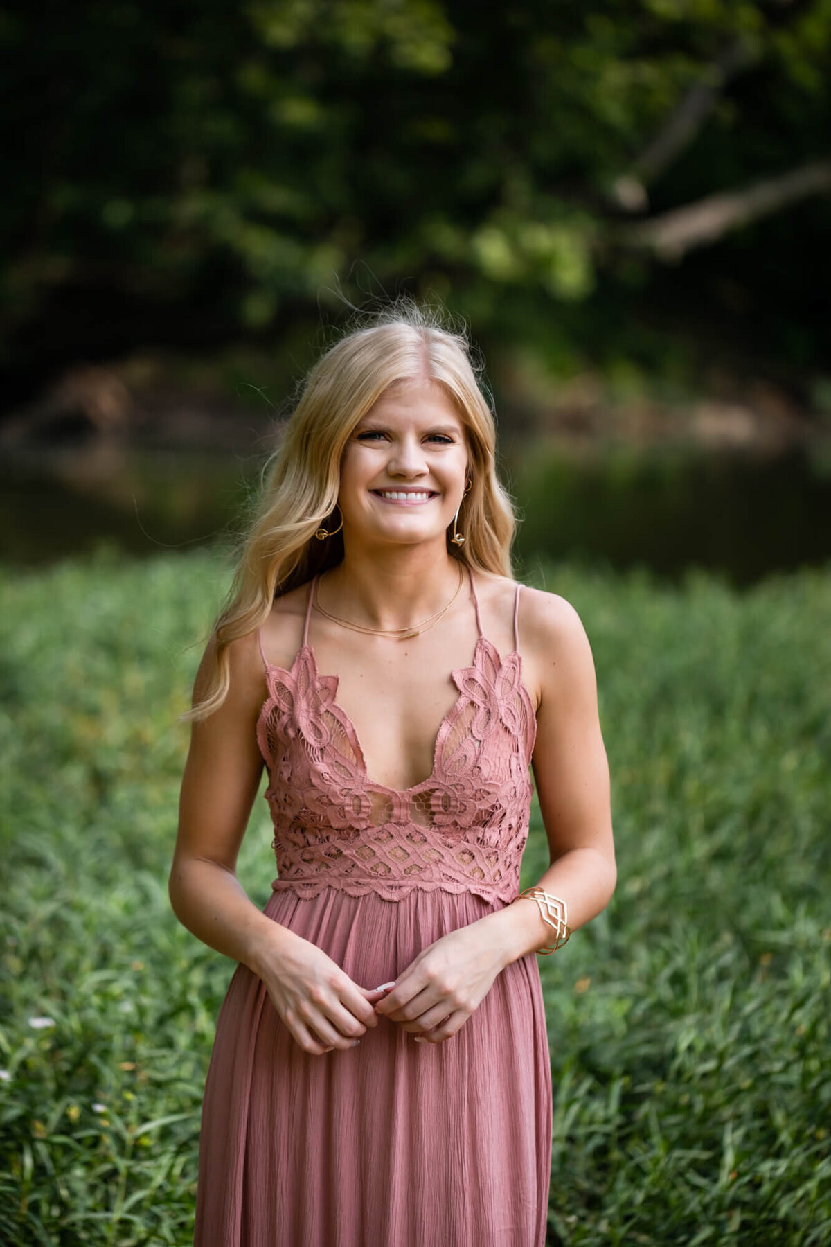 A lovely senior girl poses for portraits in a dusty pink lace dress standing in front of fresh green lake grass. Captured by Springfield, MO senior photographer Dynae Levingston.
