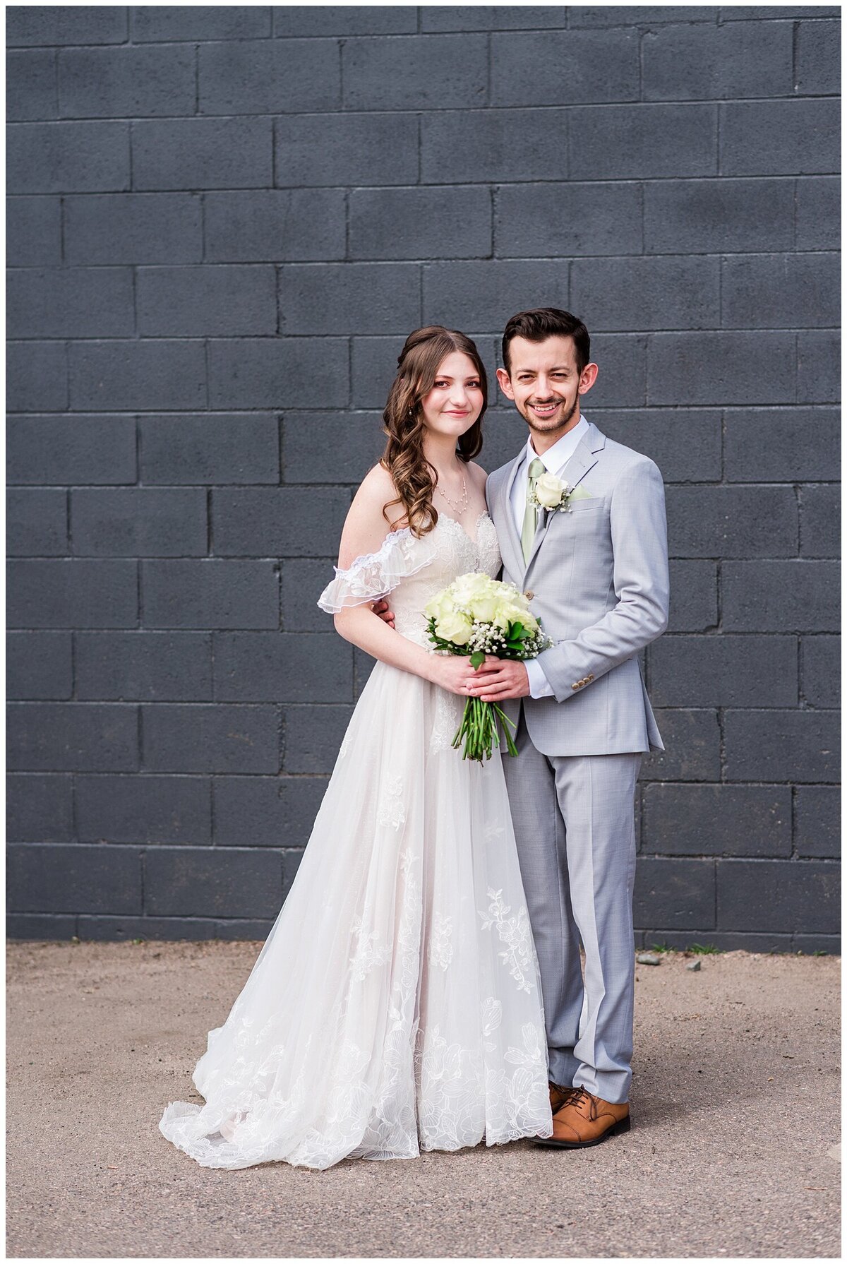 A bride and groom stand side by side next to each other holding the bride's white rose bouquet and both smile for the camera while standing in front of a black wall outside of skylight venue in downtown Denver