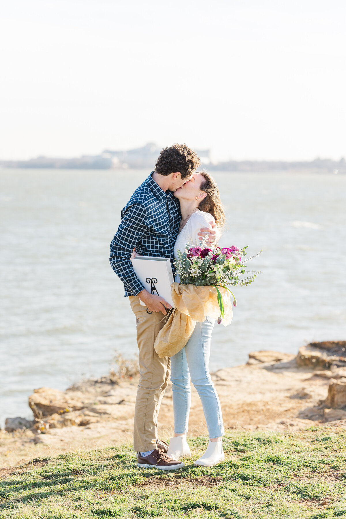 A joyful post-proposal kiss on the rocky shore of Lake Lewisville, surrounded by soft sunlight and a bouquet of fresh flowers. A natural and heartfelt moment captured in North Texas.