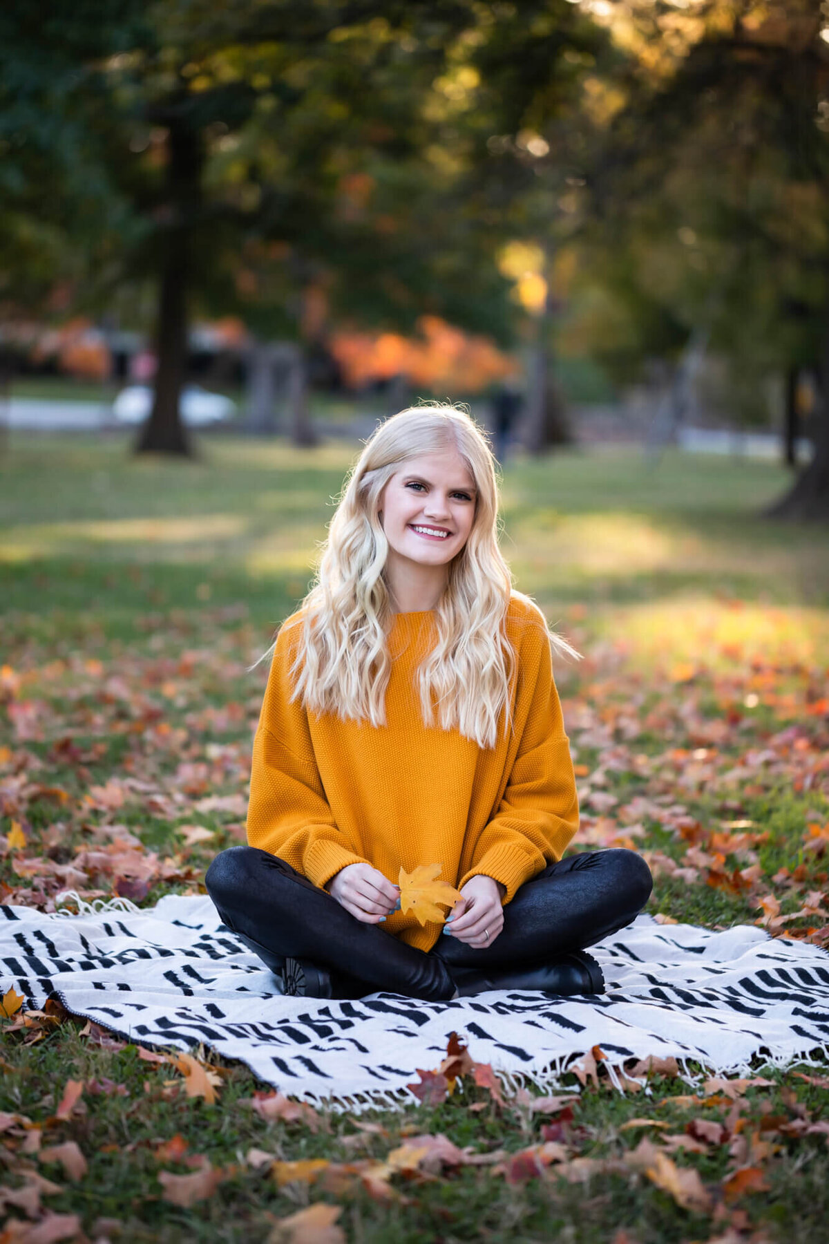 A lovely and relaxed fall senior picture of a beautiful blonde teen wearing an orange sweater and sitting cross legged on a black and white blanket surrounded by Fall leaves. Captured by Springfield, MO senior photographer Dynae Levingston.