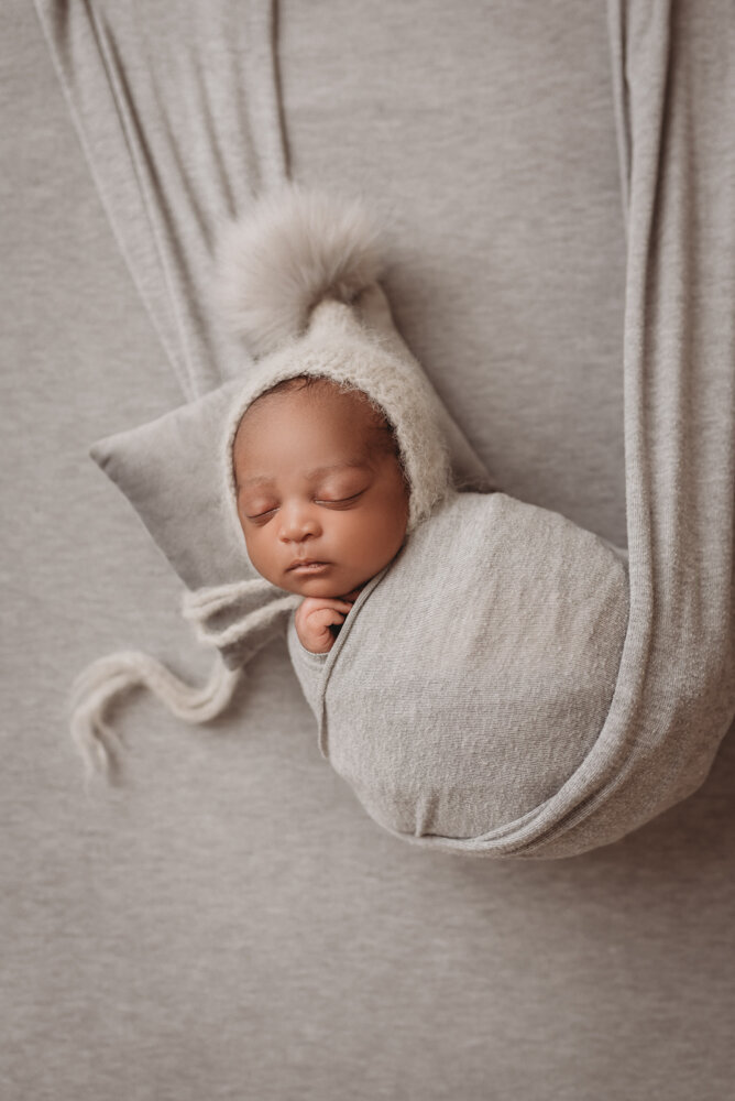 Newborn photos with Atlanta, Georgia newborn photographer of sleeping baby boy with hands under chin wrapped in gray swaddle in gray fabric swing and puff ball gray hat