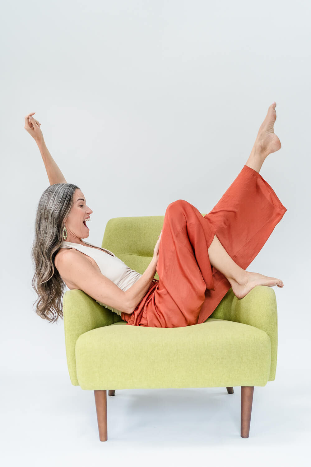 Jenn Pike playfully stretching legs up in lime green accent chair, wearing rust pants and tank.