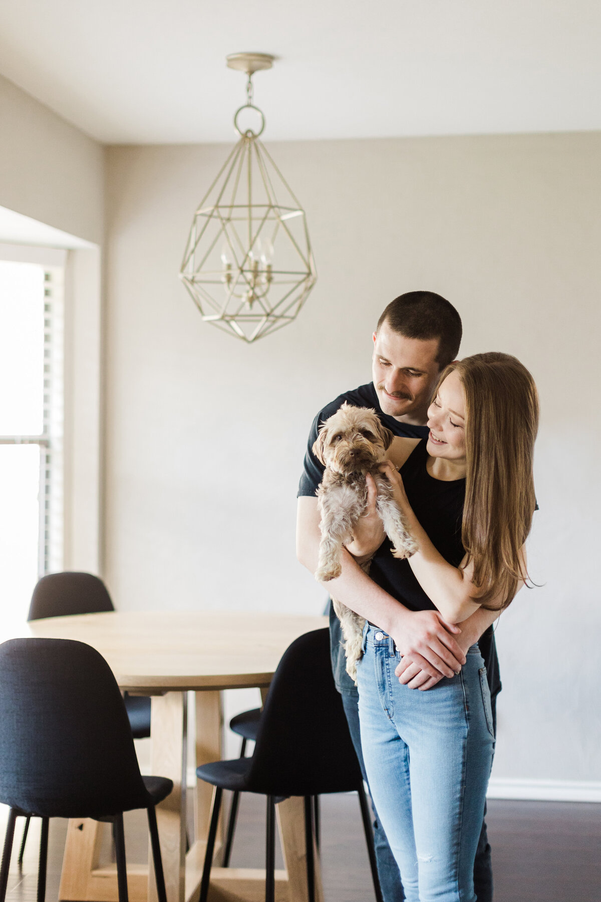 A photo of a man being the big spoon while his fiancé is both the little spoon and is holding their dog in their home during their engagement session in Fort Worth, Texas. Both the man and woman are wearing black shirts and jeans. Their dining room table can be seen in the background.
