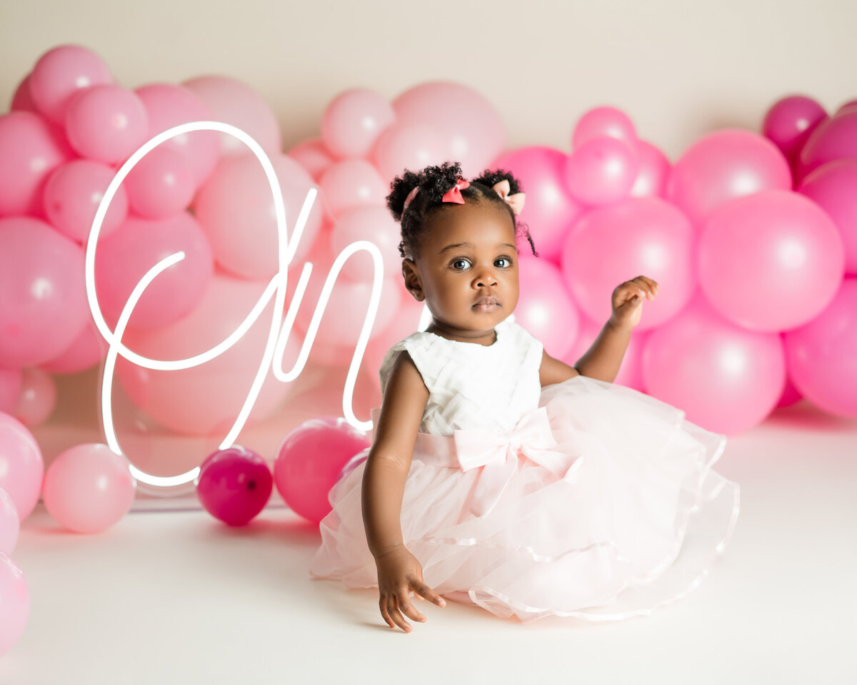 Baby girl in pink dress celebrating first birthday with cake and gold balloons by Houston photographer Lissette G