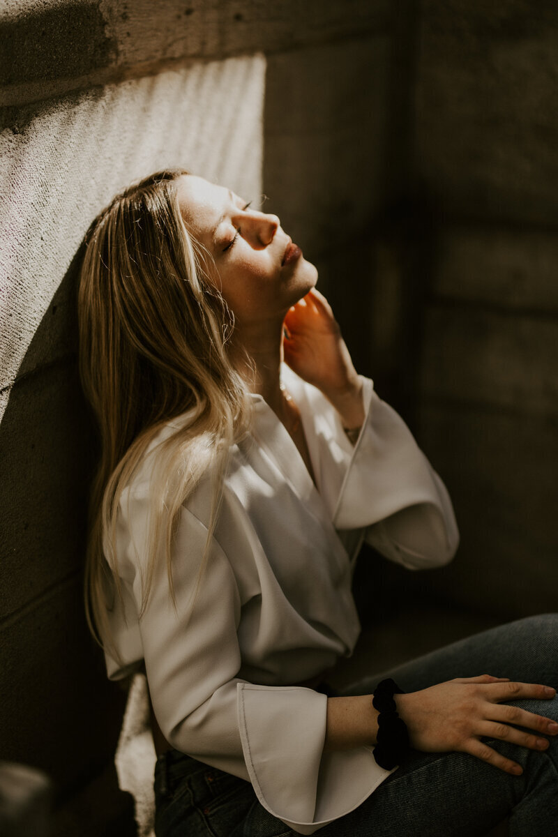 Femme blonde assise, adossée à un mur, visage en arrière et yeux fermés, inondé de lumière douce capturée par Laura lors d'une séance photo à Montréal.