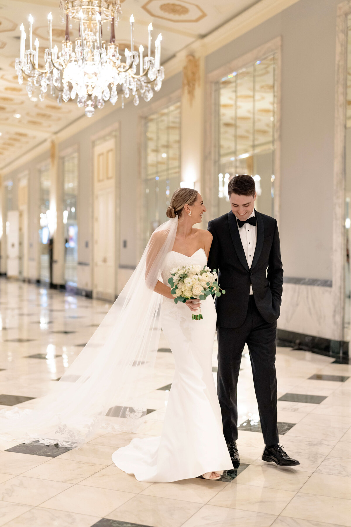 A bride and groom walk down a marble hallway. The bride wears a white dress with a long veil and holds a bouquet of white flowers. The groom wears a black tuxedo. An ornate chandelier hangs above them.