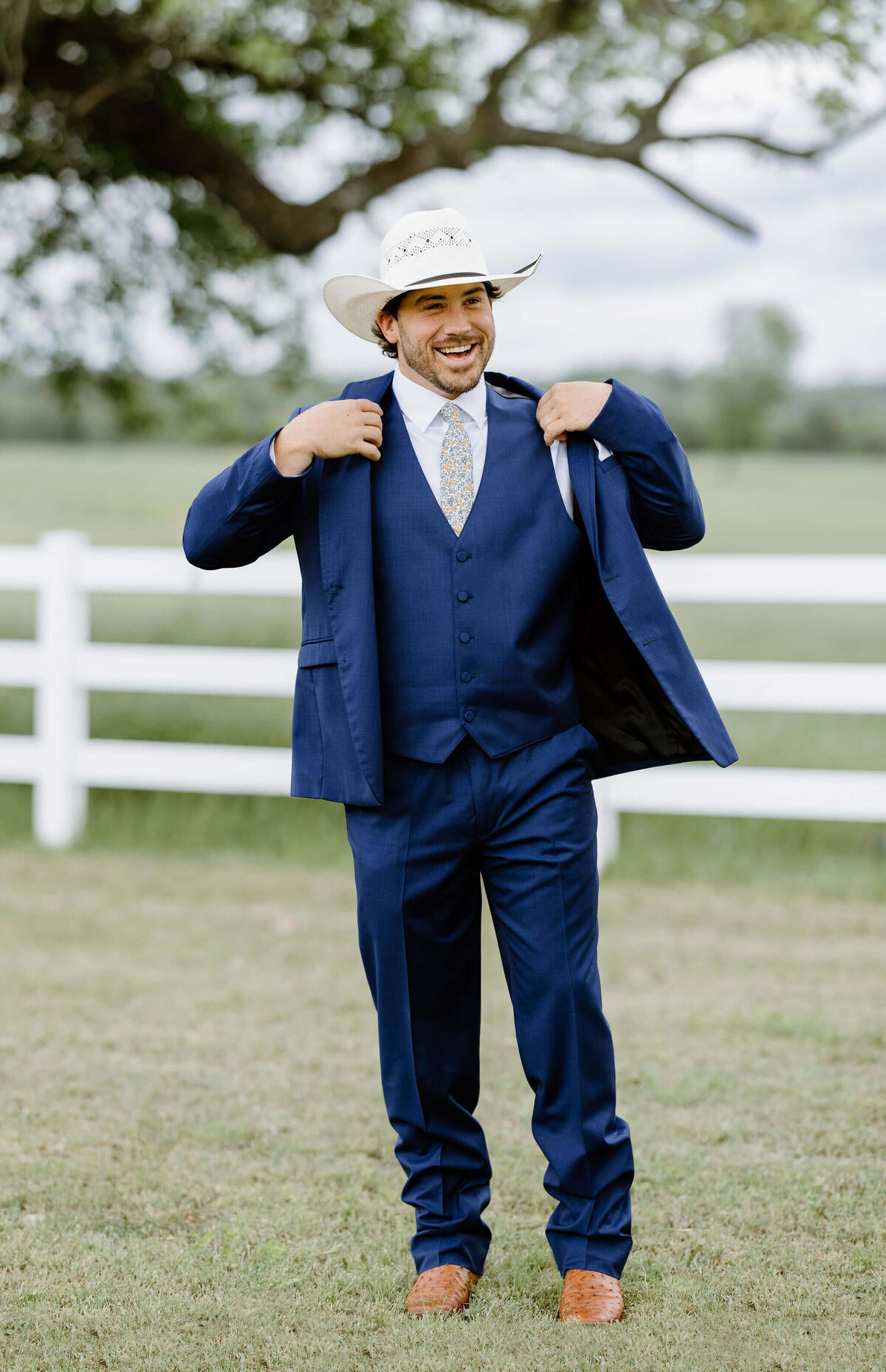 candid wedding day portrait of groom in navy suit and straw cowboy hat putting on suit coat