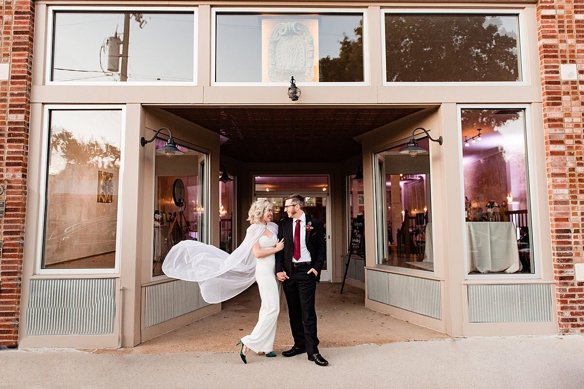 The bride and groom stand in front of a store front framed by the many windows in the brick structure. The groom looks at the bride. He is wearing a black suit with a  dark red tie. The bride is leaning into the groom as she takes his arm. The light breeze blows her cape up and out behind her.