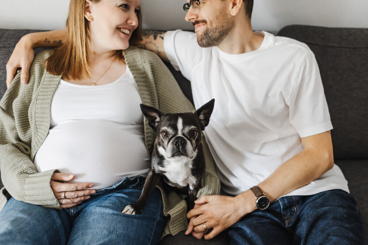 Expecting couple  sitting on couch with their pet dog. Dog is looking at the camera while the couple is looking at each other smiling.
