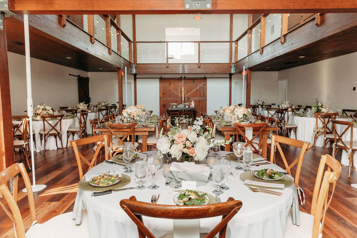 A charming Leesburg VA wedding venue showcases a rustic reception hall with round tables draped in white linens, wooden chairs, and floral centerpieces. Salads and glassware adorn each table. The room boasts a wooden floor and balcony, with natural light pouring in to create an enchanting atmosphere.