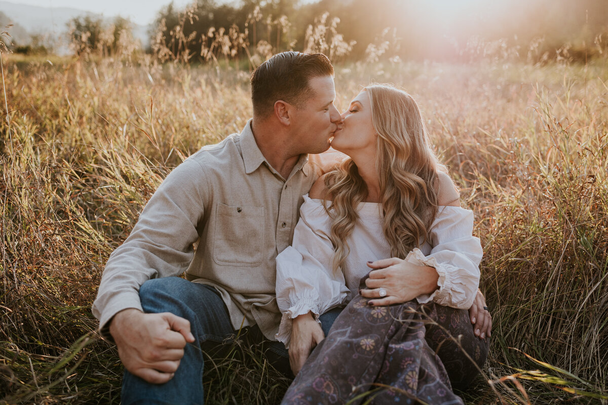 Couple sits in golden field and kiss while couddled close together.