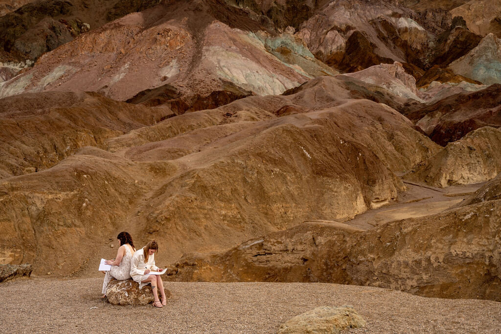 nTwo brides sitting together at Artist's Palette in Death Valley, one bride in a black dress, as they write their vows, surrounded by the vibrant, colorful hills of the desert landscape.d Canyon Elope