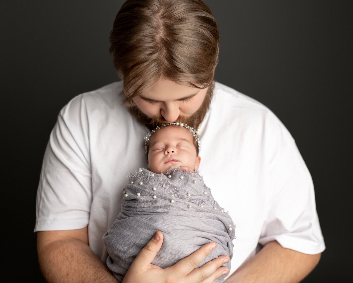 Dad kissing his newborn daughter’s forehead, swaddled in a soft gray knit wrap with pearl accents, during their studio session. Captured by Inland Empire newborn photographer Kaitlyn Dawn Photography.