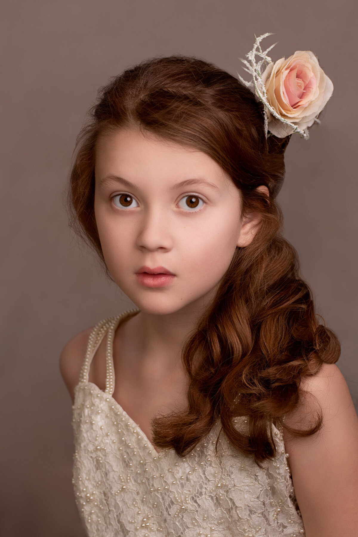 Elegant fine art portrait of a young girl wearing a lace dress with a delicate rose in her hair, posed against a soft brown backdrop. Captured in Overland Park, Kansas, this setup is ideal for families seeking classic and refined child photography.