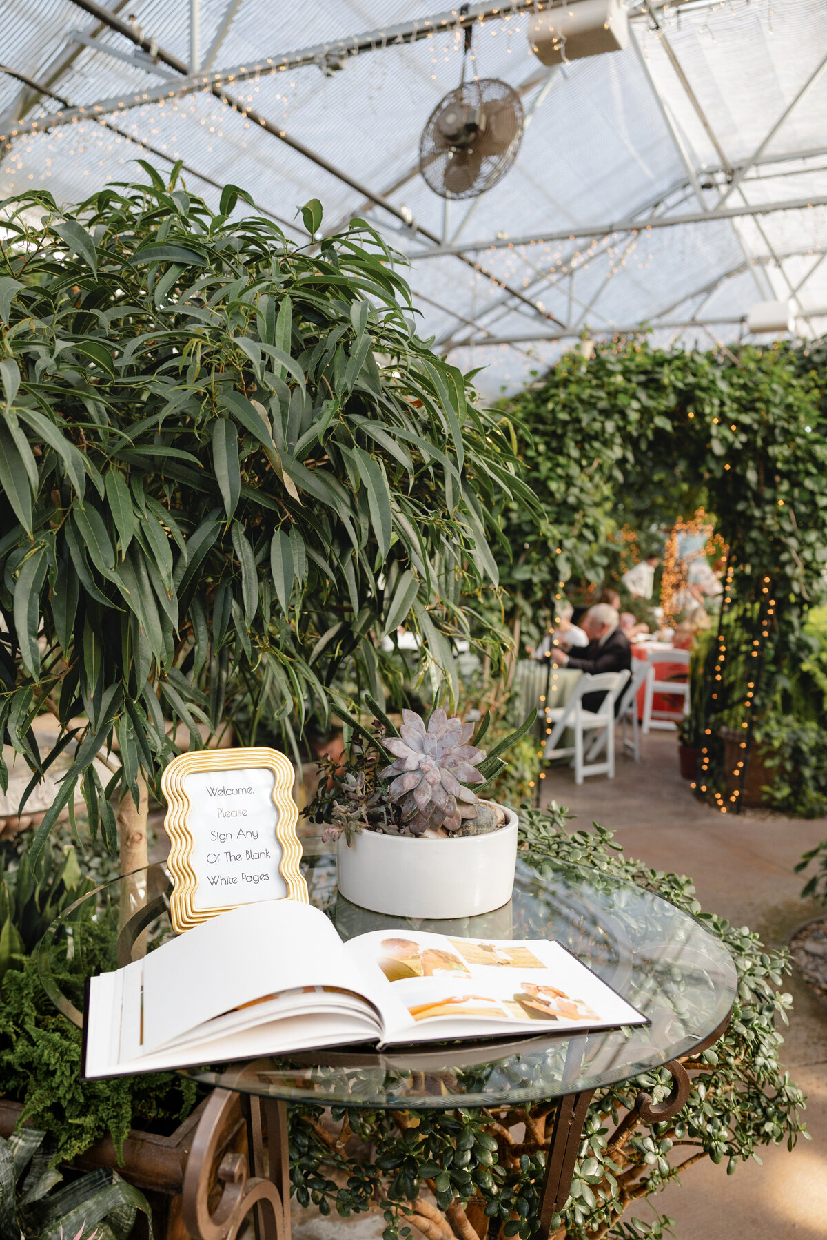 A guestbook table at the wedding reception, adorned with a potted succulent and a sign asking guests to sign the blank pages, set in a greenhouse venue with twinkling lights