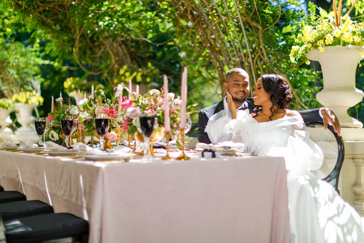 Elegant outdoor wedding tablescape at The Liriodendron Mansion featuring lush pink and green floral arrangements, gold accents, and pink taper candles, with a smiling couple seated at the head of the table under a sunlit garden canopy.