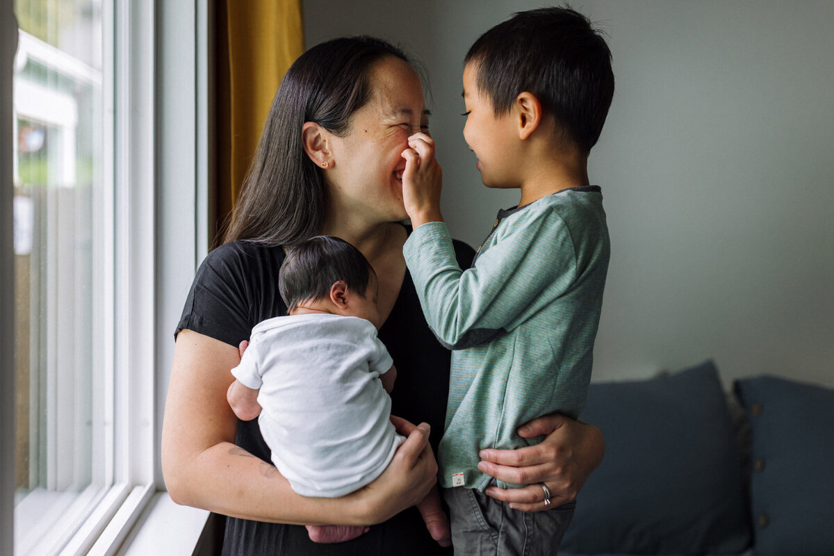 boy playing with mom's face by window while she holds newborn in white onesie