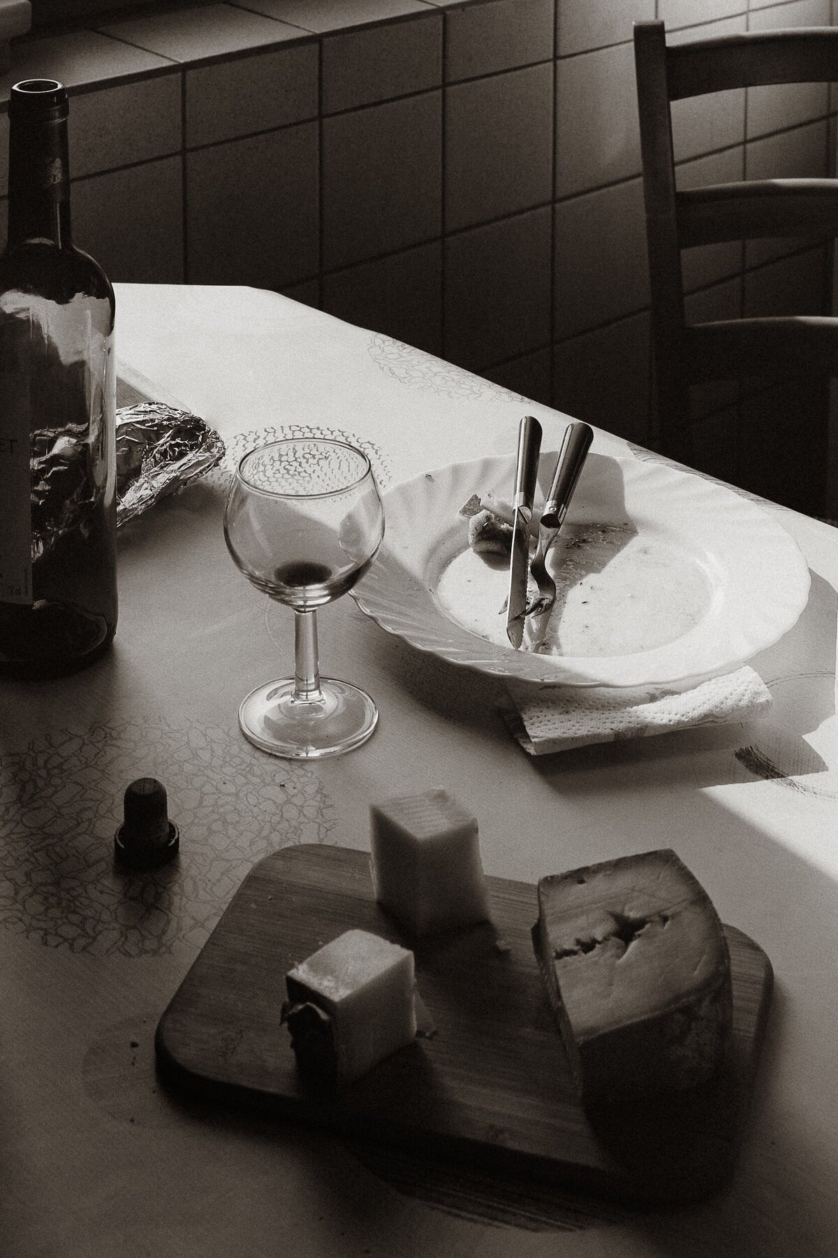 Photograph of a table after a meal, with a half eaten cheese platter, next to an empty plate, empty glass and empty bottle of wine, part of The Quiet Gift series by Marie Kenny.