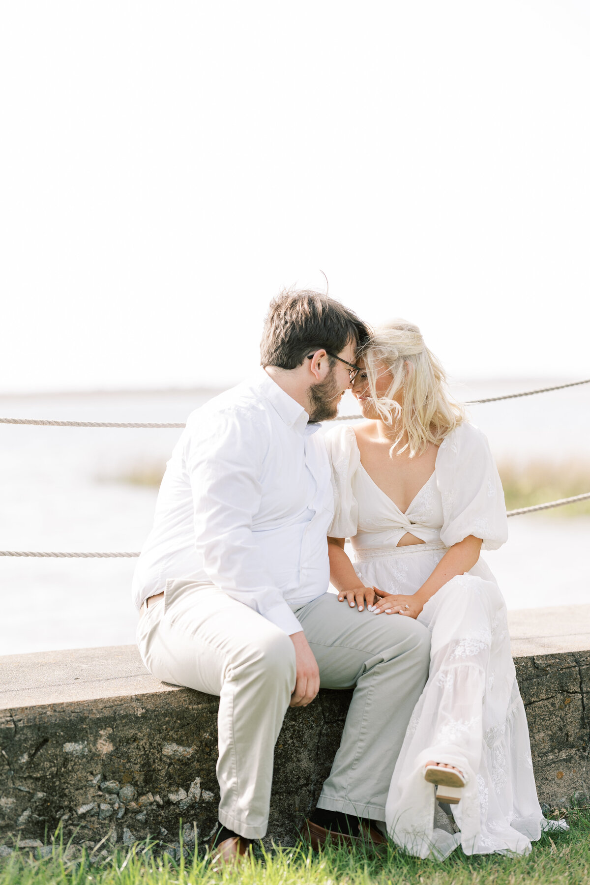 an engaged couple sitting on a partition with water in the background
