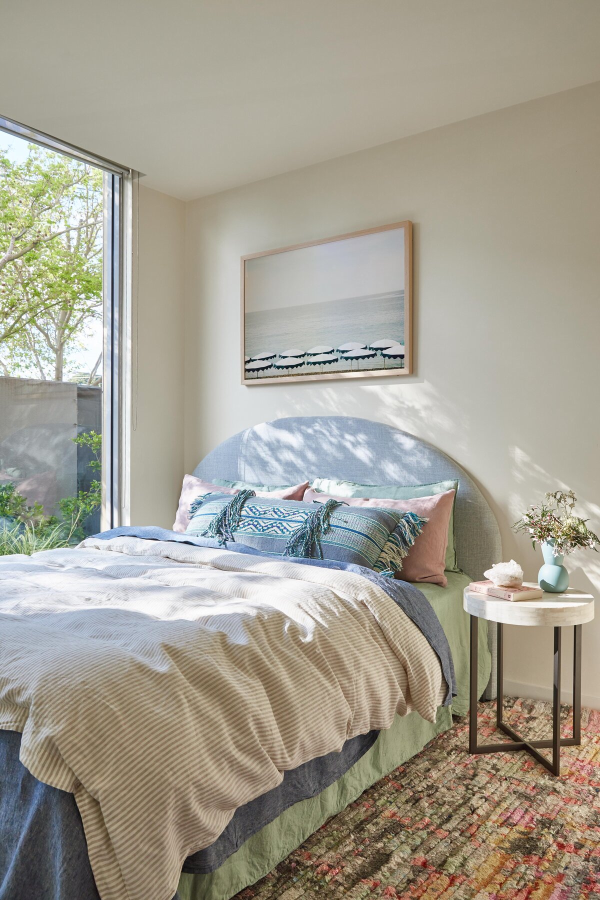 Sunlit Elwood bedroom with a curved upholstered headboard, seascape print above the bed and layered linen bedding; marble side table and garden outlook add softness.