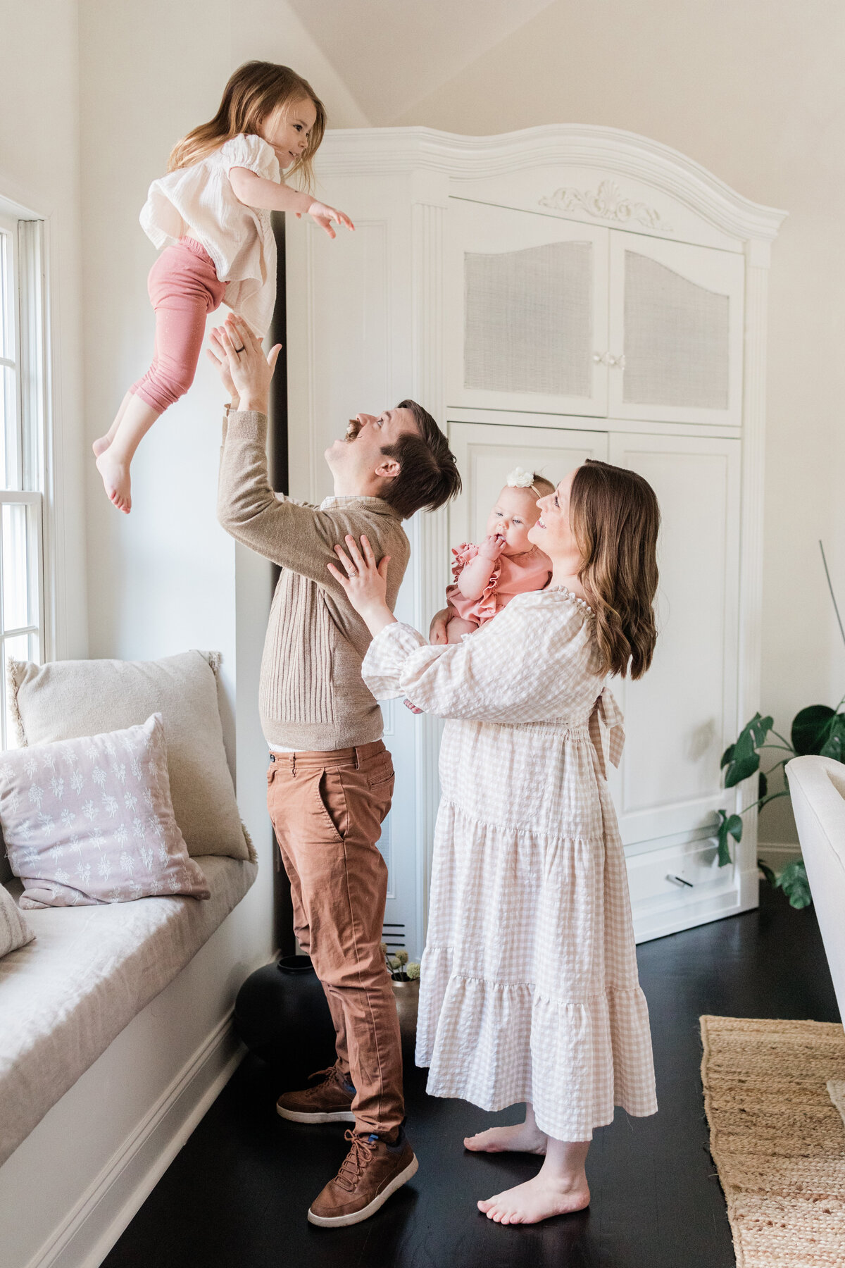 Mom holding baby and dad tossing toddler into air in front of window