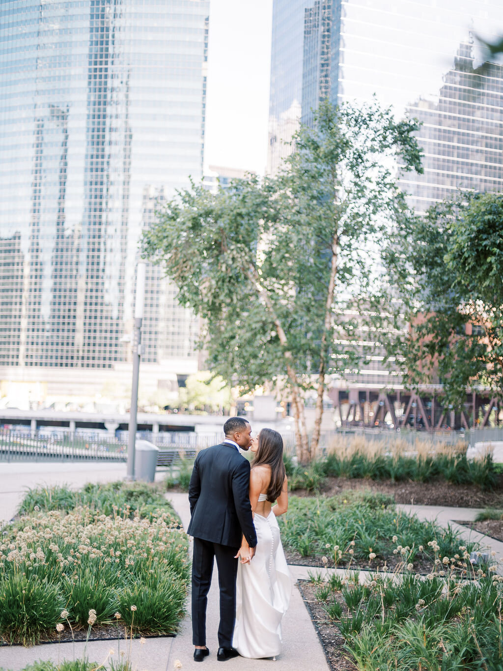 The couple walks hand in hand through a lush garden in Chicago, with the city skyline in the background.