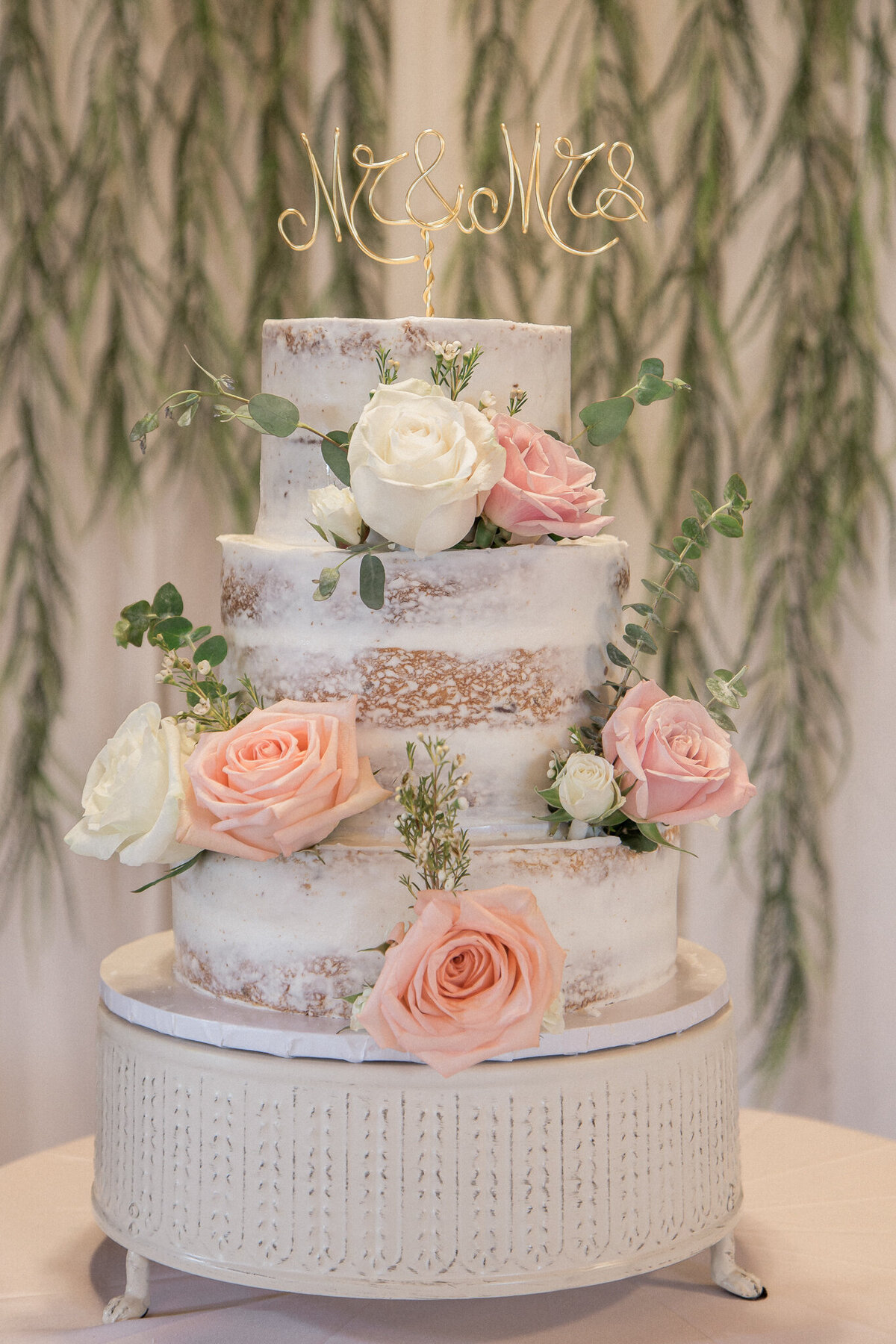The three tiered wedding cake, topped with Mr & Mrs topper is decorated with white and pink roses in front of strands of eucalyptus leaves