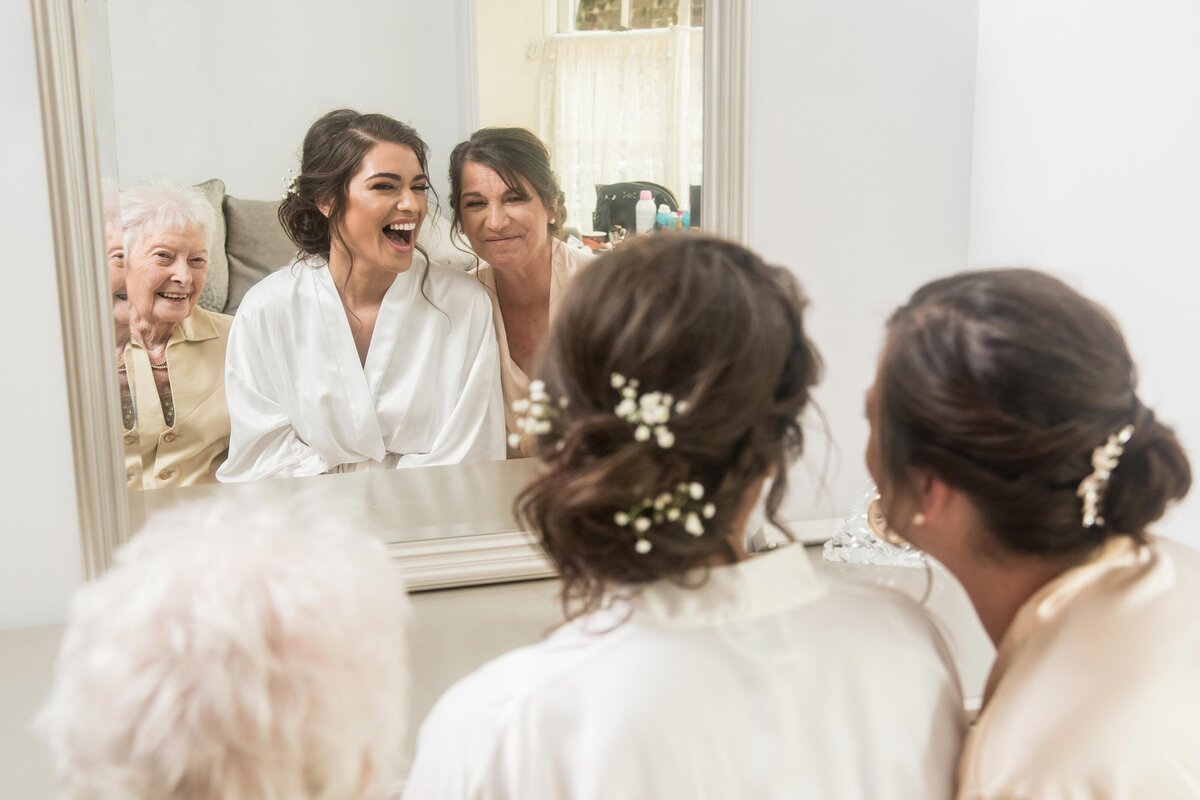 a bride and her mother and grandmother share a moment in front of a mirror