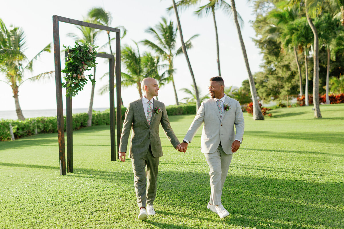 A newlywed couple in suits holding hands and walking in a field 