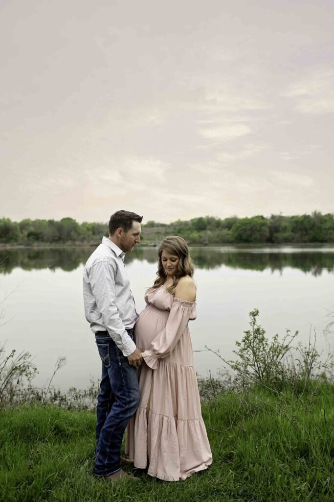 A serene maternity photo of the expectant mom standing by the lake, gently cradling her belly as the soft water reflects the clear sky, captured by Chunky Monkey Photography.