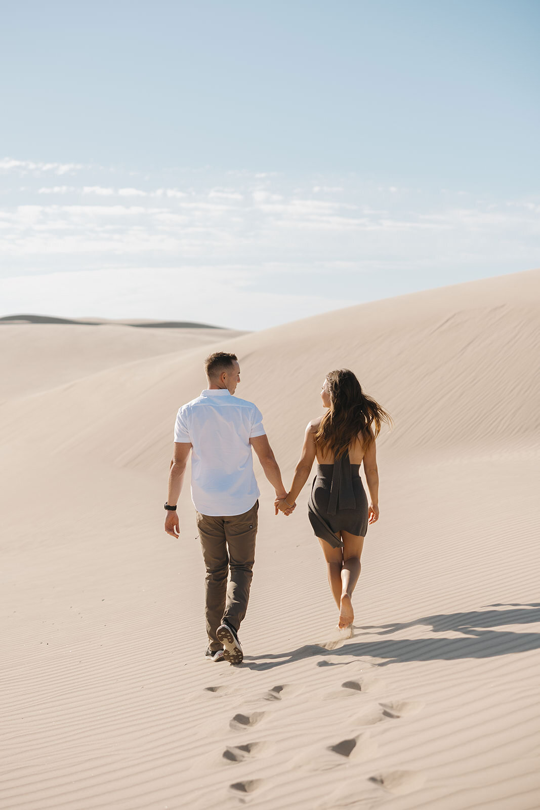 Engaged couple walking hand in hand across sand dunes under a blue sky